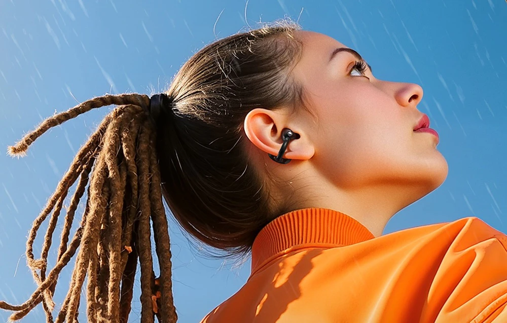 Femeie cu dreadlocks și cască OpenRock în hanorac portocaliu pe fundal de cer