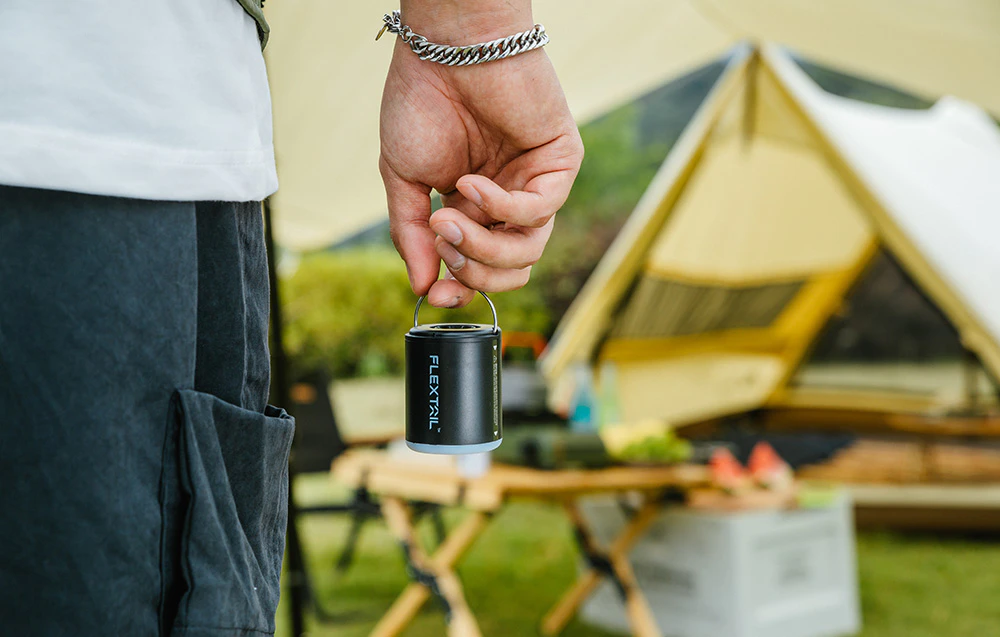 Prim-plan al unei mâini care ține pompa compactă Flextail de mânerul metalic, pe fundalul unui cort și al unei mese de camping