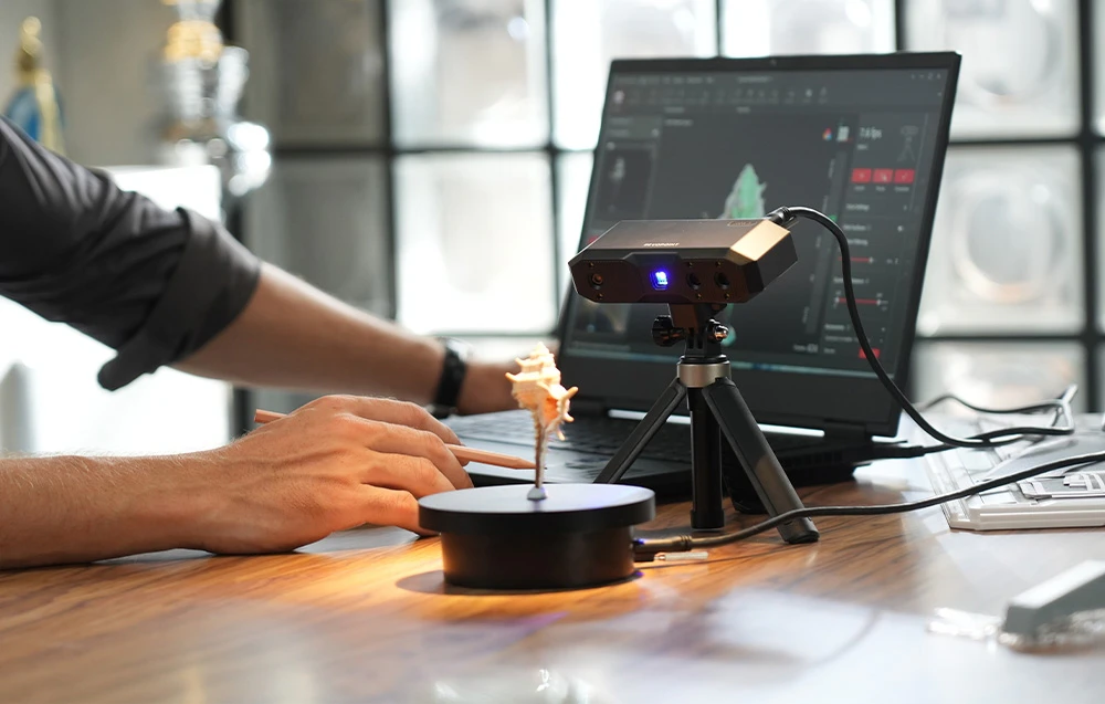 Scanerul 3D Revopoint Mini 2 scanând o scoică pe o masă rotativă, lângă un laptop care afișează modelul 3D în software