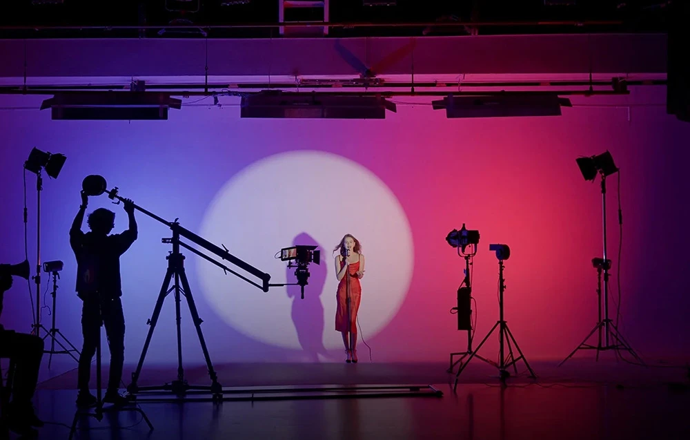 O femeie cântă pe scenă în lumina unui spot mare, pe fundalul unui perete roz-violet.