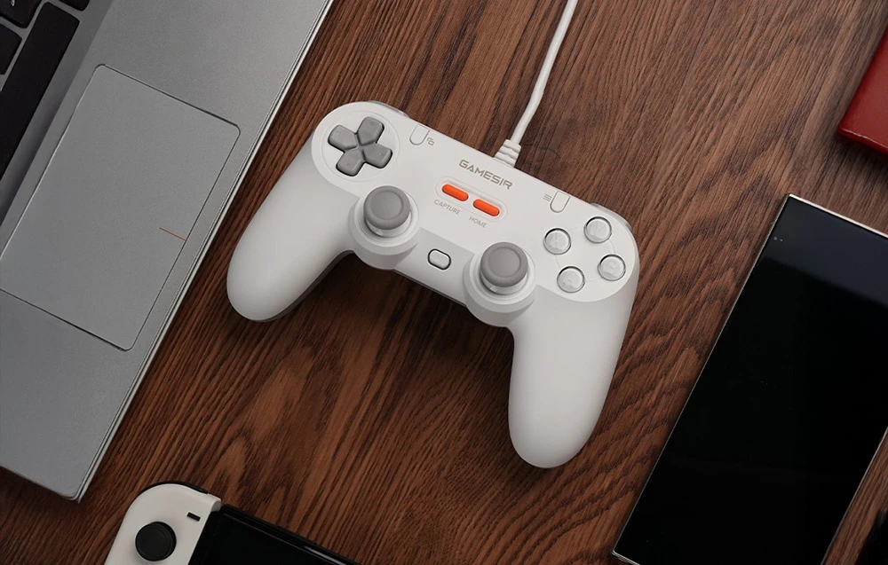 Un controller GameSir alb conectat cu un cablu, așezat pe o masă de lemn lângă un laptop și smartphone-uri.