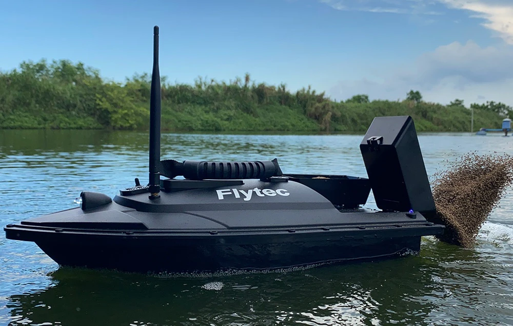 Barca de momeală Flytec cu compartiment pentru momeală plutind pe un lac, cu un mal verde în fundal