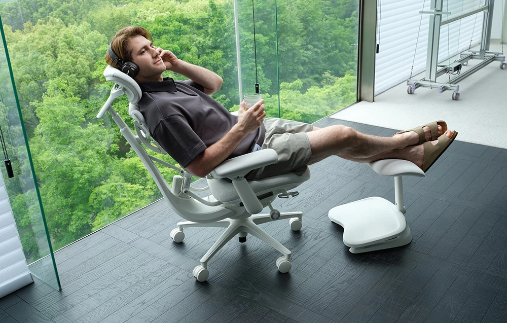 Un bărbat care poartă căști se relaxează într-un scaun ergonomic alb cu suport pentru picioare, ținând în mână un pahar cu o băutură
