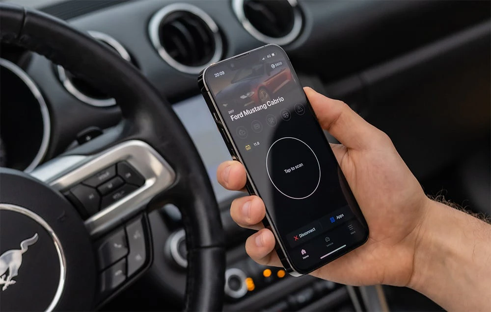 Mână care ține un smartphone cu aplicația Carly care afișează datele Ford Mustang Convertible în interiorul mașinii