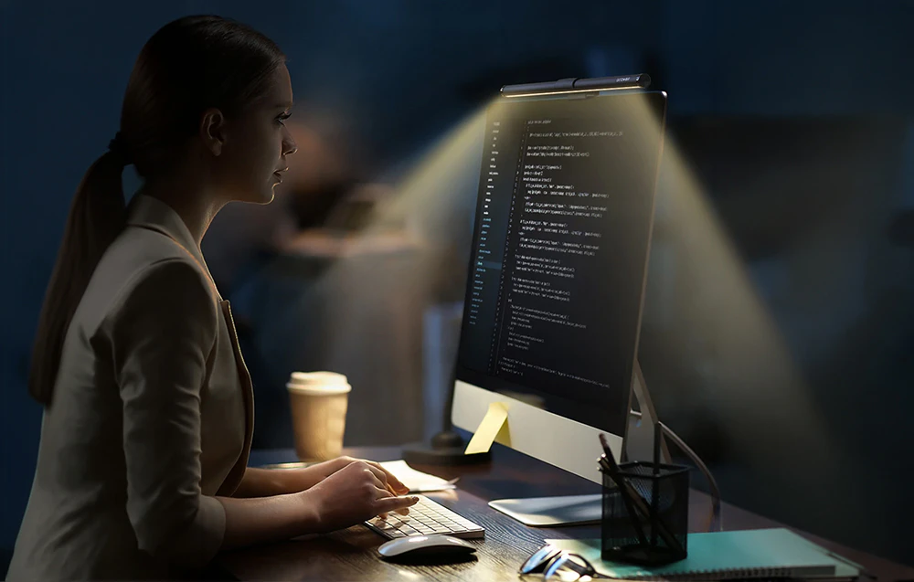 O femeie așezată la birou, tastând pe o tastatură în timp ce se uită la un monitor cu o lampă atașată