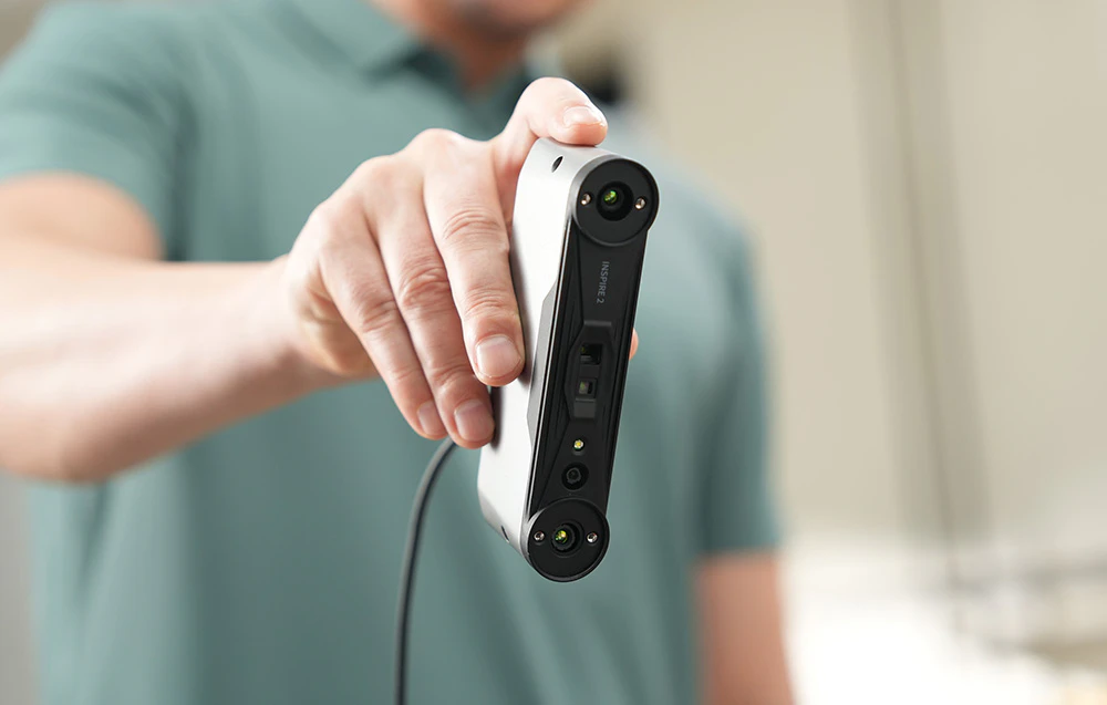 Un bărbat îmbrăcat într-o cămașă verde ține în mână un scaner 3D Inspire 2 argintiu, poziționat vertical