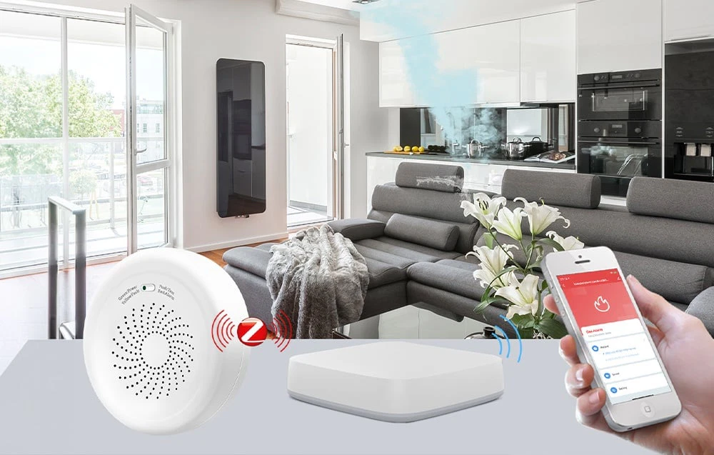 Un detector de gaz, un gateway ZigBee și un smartphone cu aplicația pe fundalul unui living cu o canapea gri și o bucătărie albă