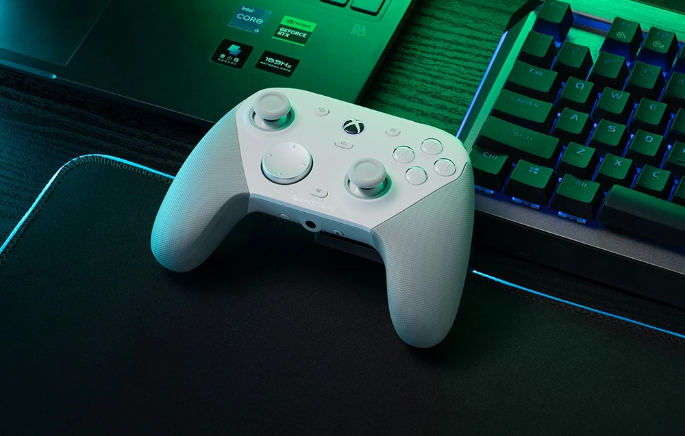 Un controler GameSir alb așezat pe un pad lângă un laptop și o tastatură.