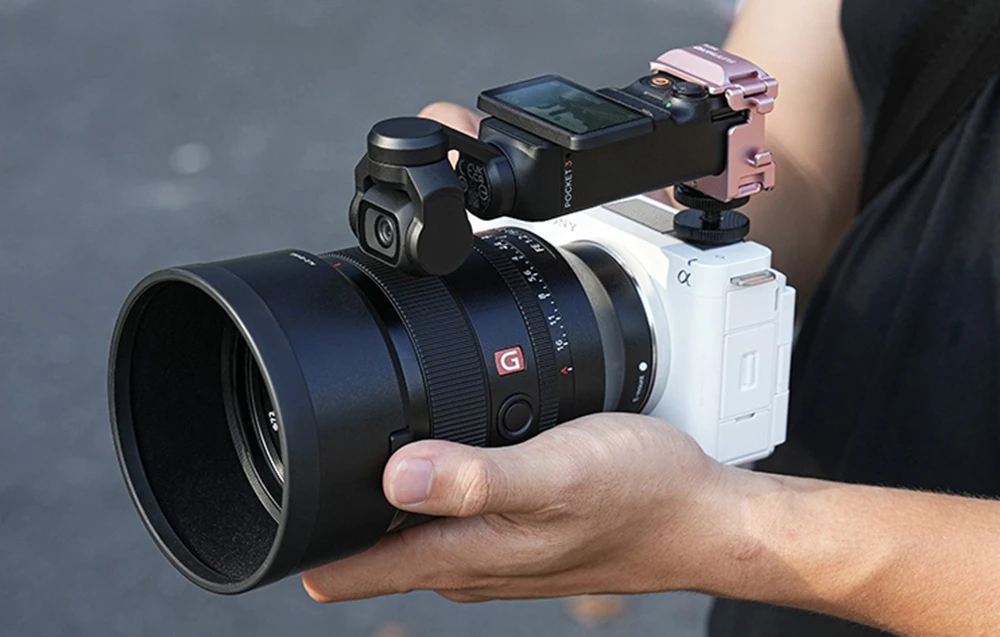 O cameră DJI Pocket în montură roz montată deasupra unei camere Sony cu obiectiv mare, ținută în mână