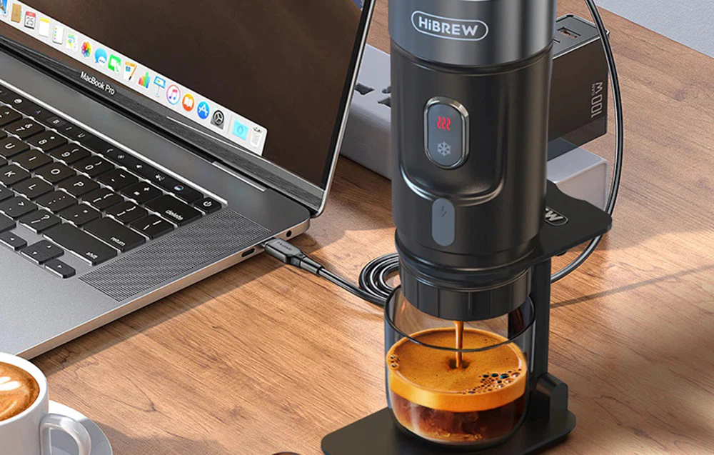 Aparatul de cafea negru prepară cafea într-un pahar, fiind așezat pe un birou lângă un laptop.