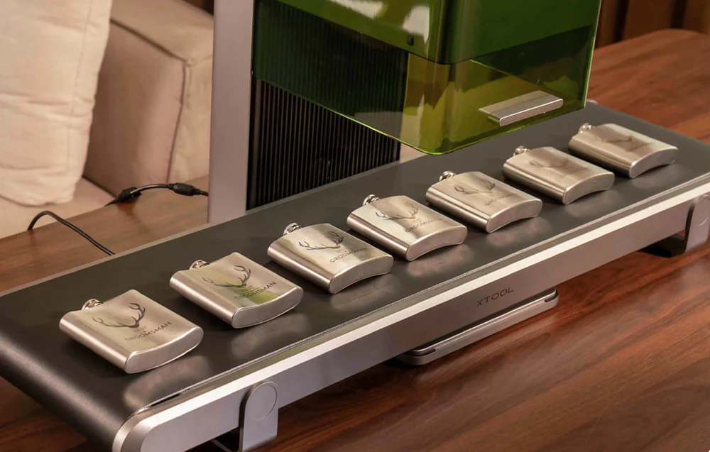 O serie de flacoane metalice gravate cu coarne de cerb, aranjate pe alimentatorul mobil al laserului xTool
