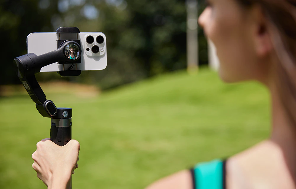O femeie se filmează în aer liber, ținând gimbalul cu smartphone-ul în mână în timp ce înregistrează.