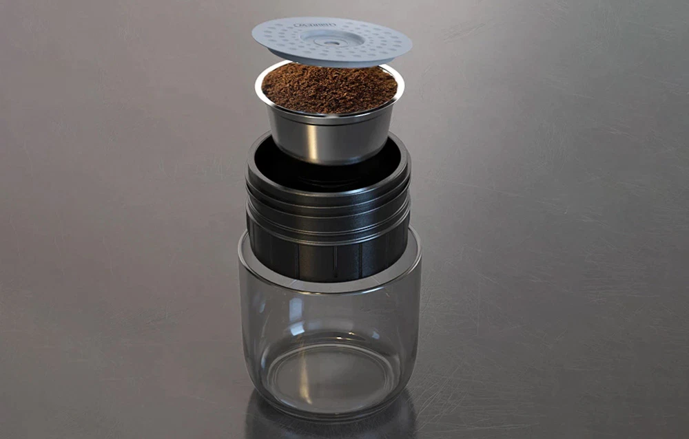Un recipient metalic cu cafea măcinată se află deasupra părții negre a aparatului de cafea și a paharului.