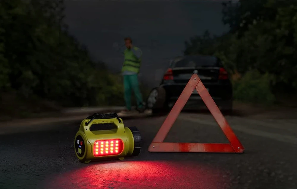 Un triunghi de avertizare lângă o lampă galbenă cu lumină roșie pe drum, o siluetă în vestă în depărtare.