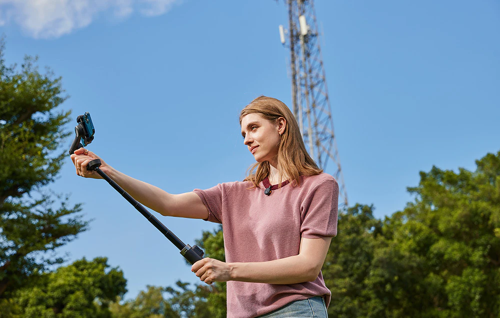 O femeie își face un selfie folosind un gimbal cu mâner extensibil, având ca fundal vegetația și cerul.