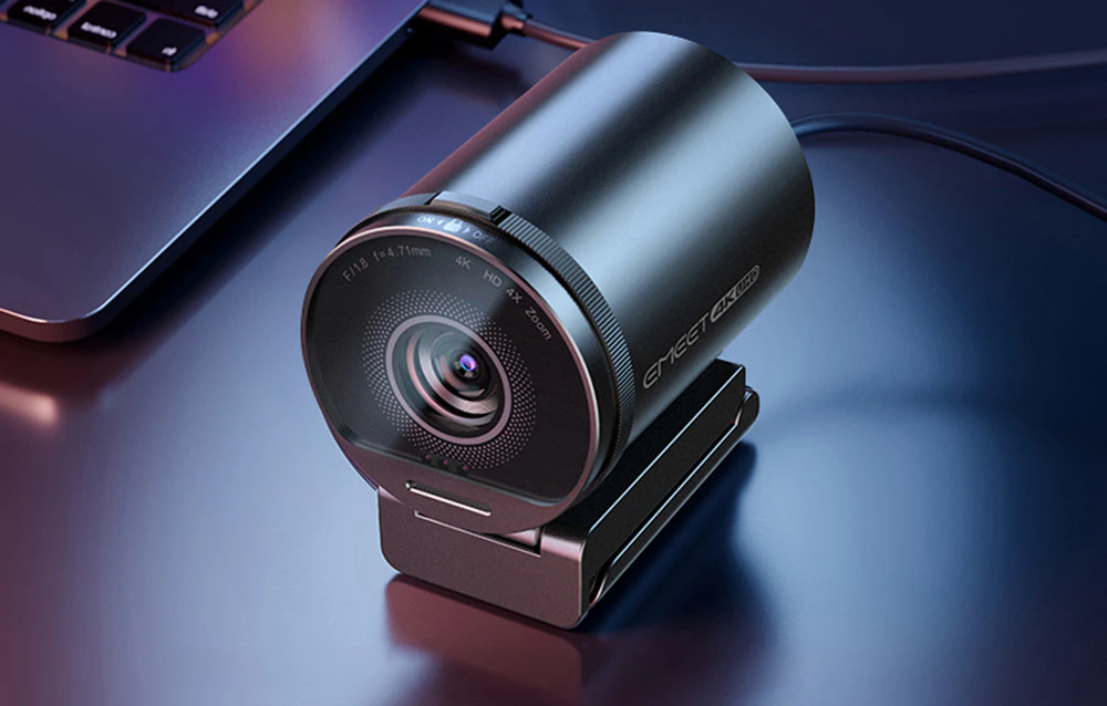 O cameră într-o carcasă metalică de culoare închisă, așezată pe tejghea lângă laptop. Aceasta poartă inscripția Emeet 4K UHD.