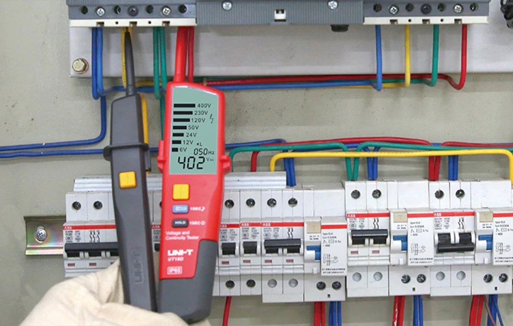 O mână înmănușată ține un tester cu afișaj digital, măsurând parametrii într-un tablou electric.