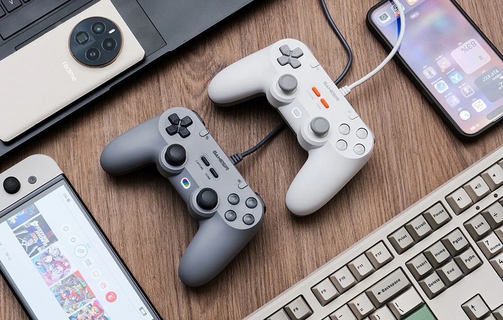 Două controlere GameSir, alb și gri, așezate pe un birou din lemn printre diverse dispozitive electronice.