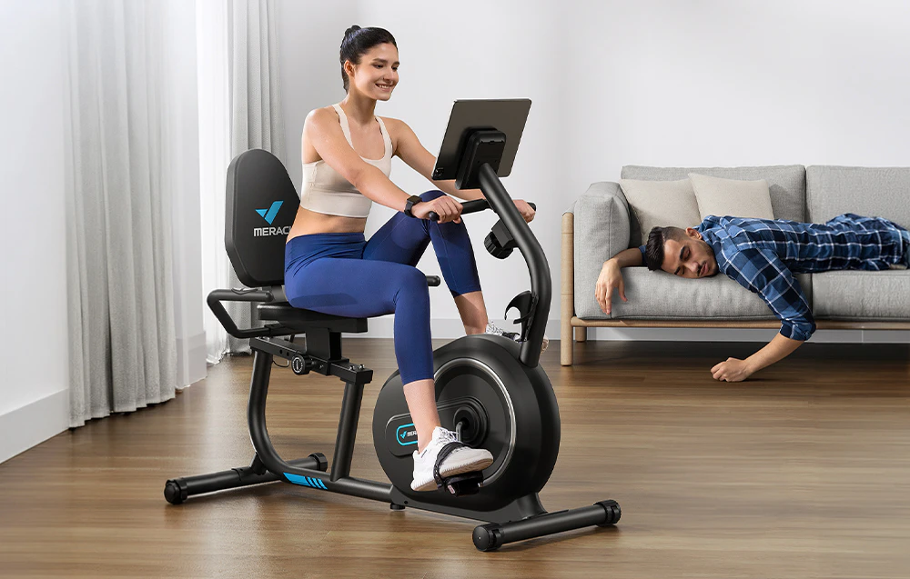 Femeie care face exerciții pe o bicicletă reclinată, bărbat care doarme pe canapeaua de lângă ea