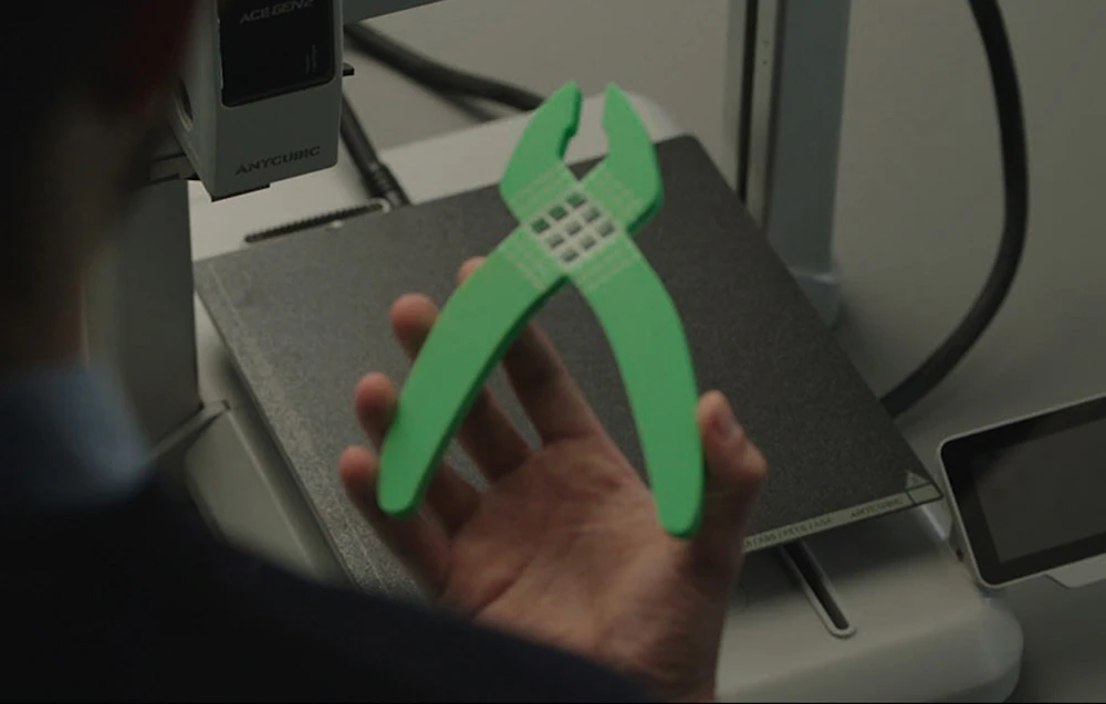 Un bărbat ține o clește verde, imprimată, deasupra platformei imprimantei 3D Anycubic