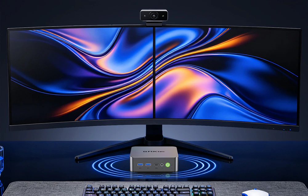 Un monitor cu ecran divizat lat, o cameră web pe marginea superioară și un mini-computer GMKtec poziționat sub ecran, lângă o tastatură și un mouse.