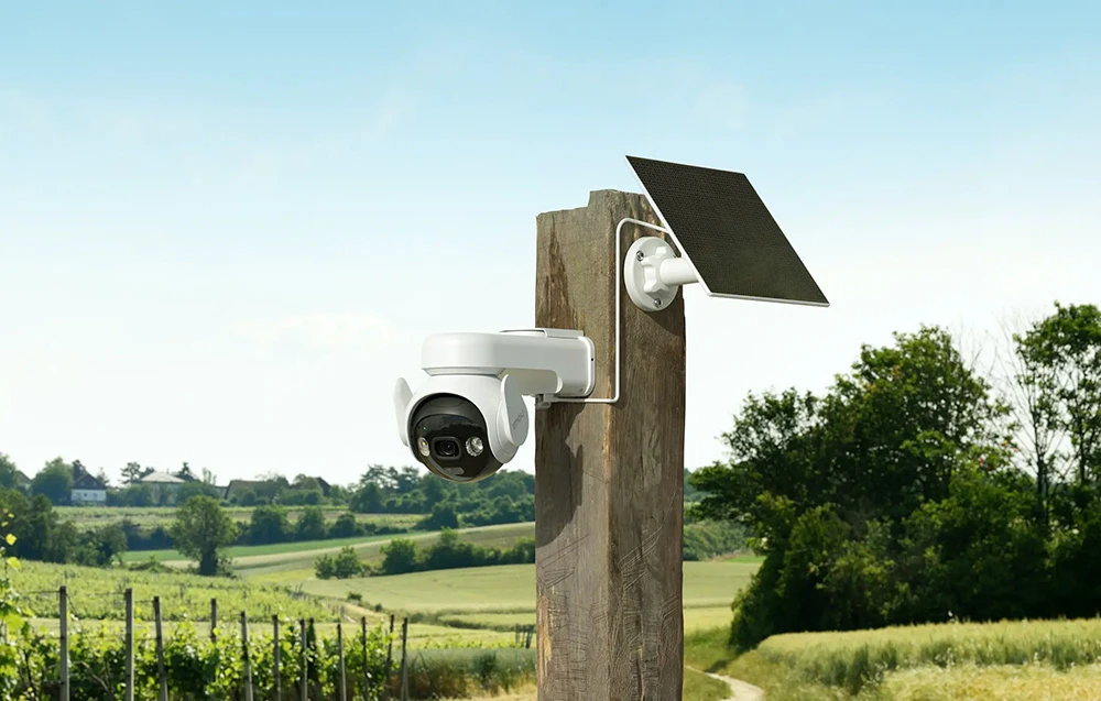 Cameră albă cu un panou solar montată pe un stâlp de lemn, pe fundalul câmpurilor, al viilor și al cerului albastru