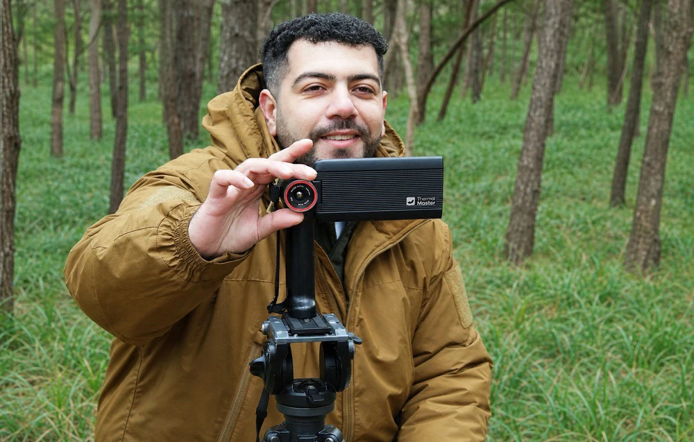 Un bărbat în pădure ține o cameră cu imagistică termică montată pe un trepied.