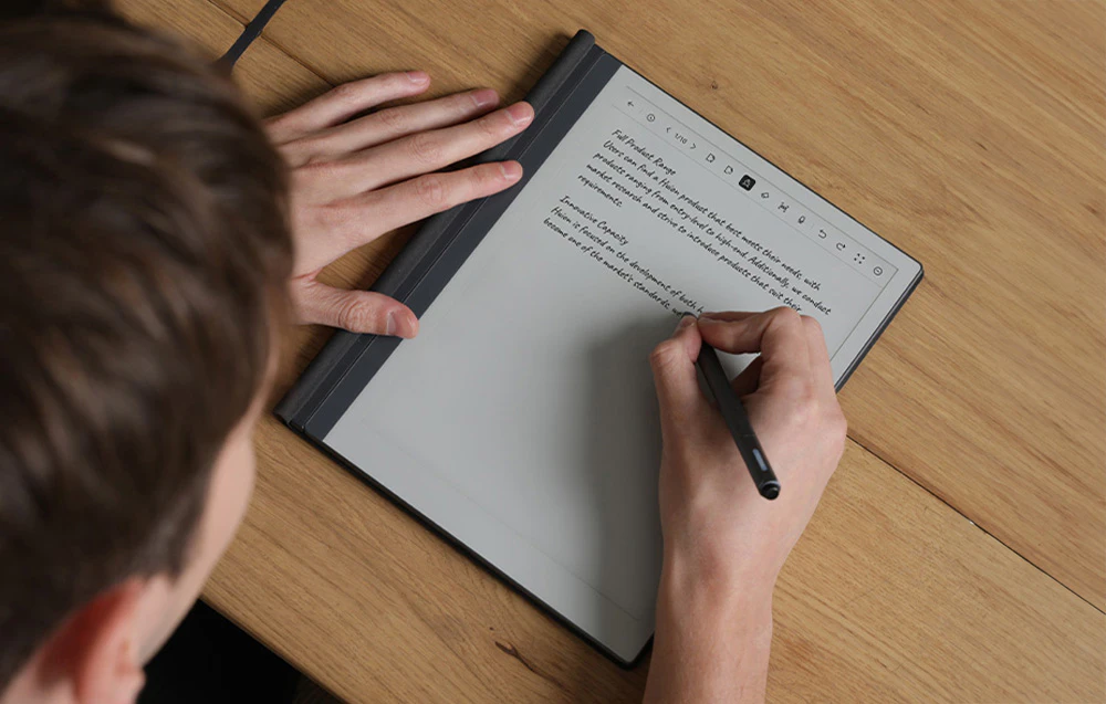 O persoană scrie cu stiloul pe ecranul unei tablete e-paper în timp ce ține dispozitivul pe o masă de lemn.