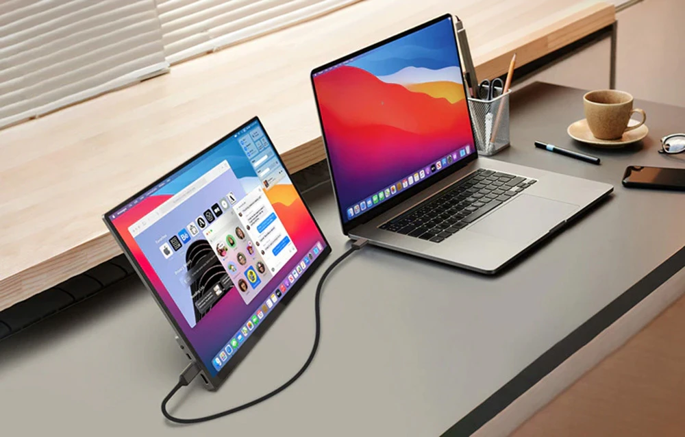 Ecranul portabil stă pe un birou lângă un laptop și servește ca monitor suplimentar conectat printr-un cablu.