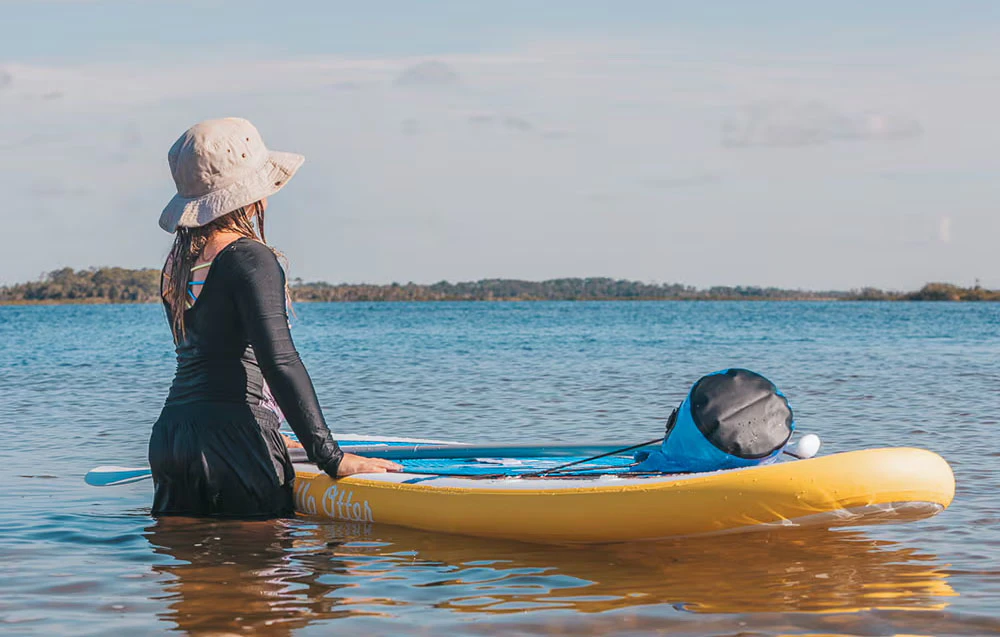 Fată cu pălărie gri stând pe apă lângă o placă de surf și o vâslă