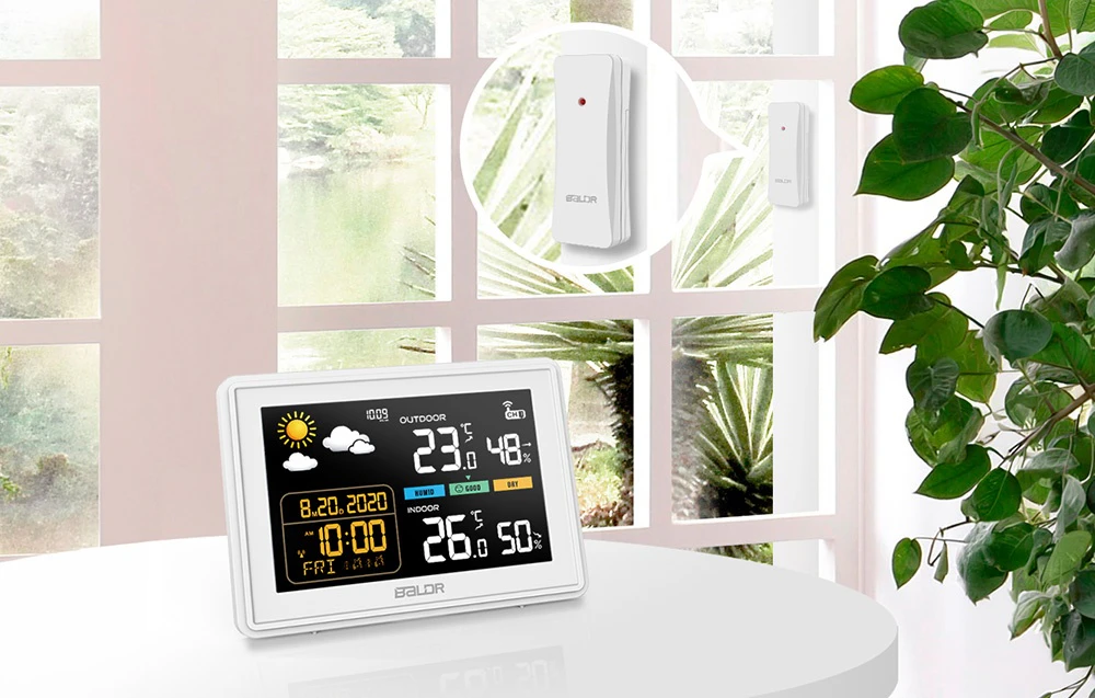 Stația meteorologică digitală Baldr se află pe o masă albă într-o cameră luminoasă. În fundal, pe fereastră, se vede un senzor exterior alb, fără fir, cu un LED roșu