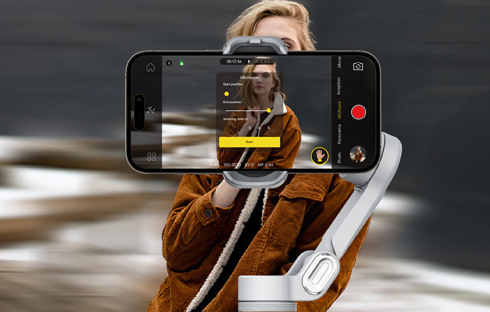 Un gimbal cu un smartphone care afișează interfața în timp ce filmează o femeie într-o jachetă maro.