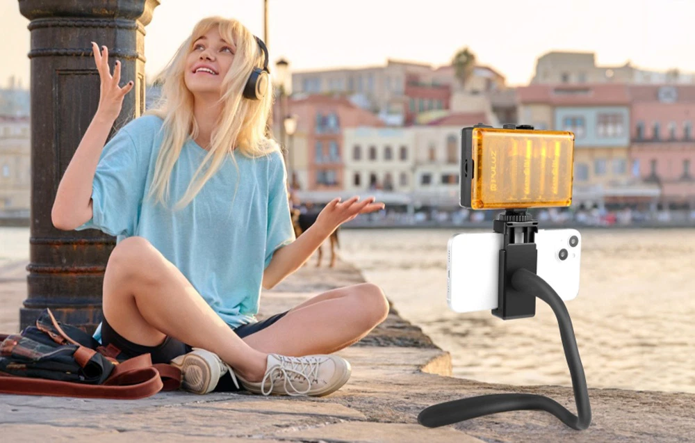 O femeie așezată pe malul apei, purtând căști, cu un smartphone așezat într-un suport cu lumină pentru înregistrare lângă ea, pe fundalul clădirilor orașului.