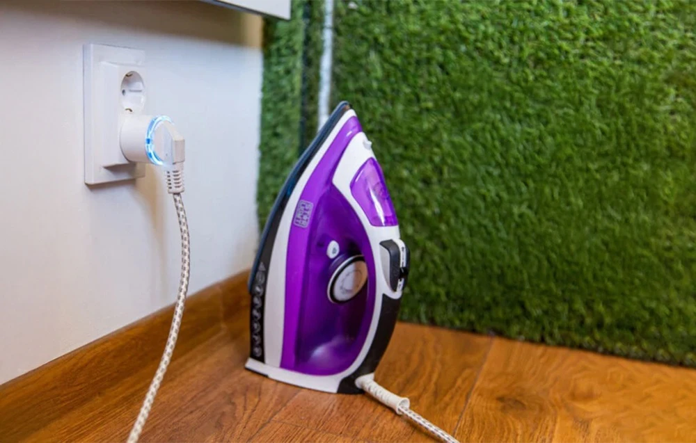 Shelly Plug S cu iluminare albastră conectat la un fier de călcat lângă un perete de iarbă artificială