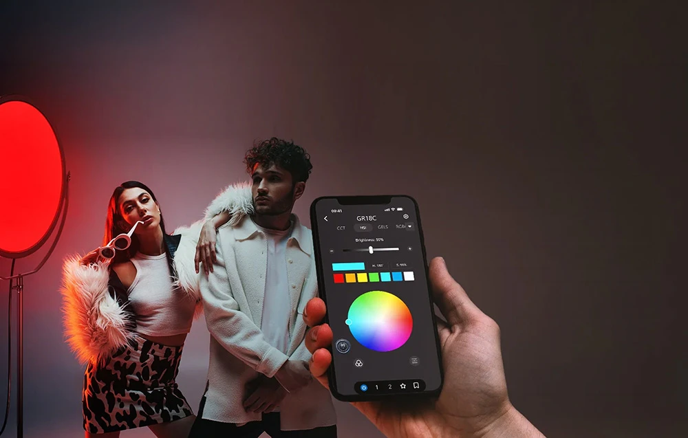 O mână ține un telefon pe care este afișată o roată de culori pentru a controla iluminarea celor două modele din fundal.