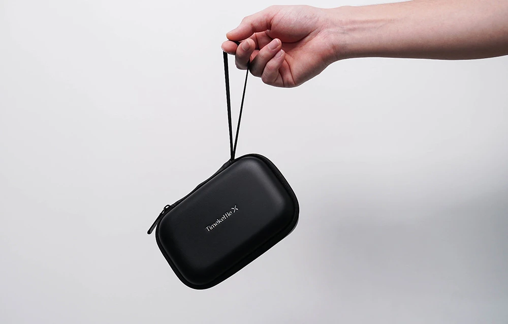 O husă de călătorie neagră, închisă, cu inscripția „Timekettle X”, care se ține în mână cu ajutorul unei curele subțiri.