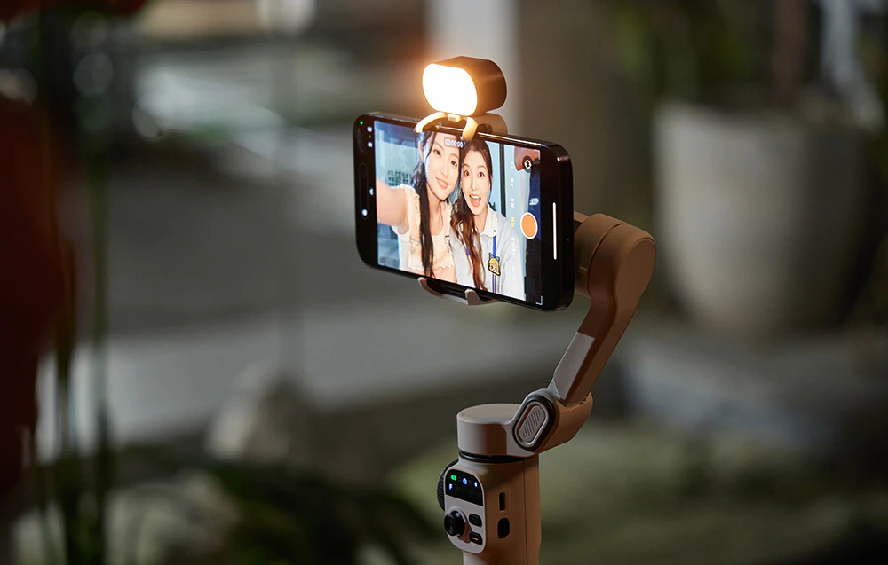 Un smartphone montat într-un gimbal bej cu lumina aprinsă, afișând un selfie cu două femei.