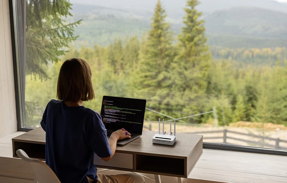 O femeie stă cu spatele la cameră la un birou, lucrând la un laptop; lângă ea se află un router alb, iar pe fereastră se vede o pădure de conifere.