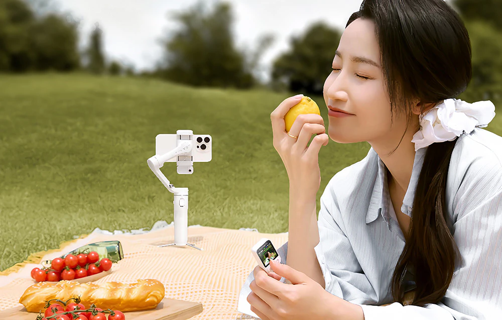 O femeie așezată pe o pătură de picnic, ținând în mână o lămâie și un smartphone, lângă un gimbal alb cu un telefon.