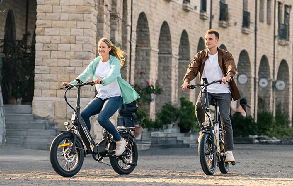 O femeie și un bărbat se deplasează cu biciclete electrice într-o piață înconjurată de clădiri din piatră