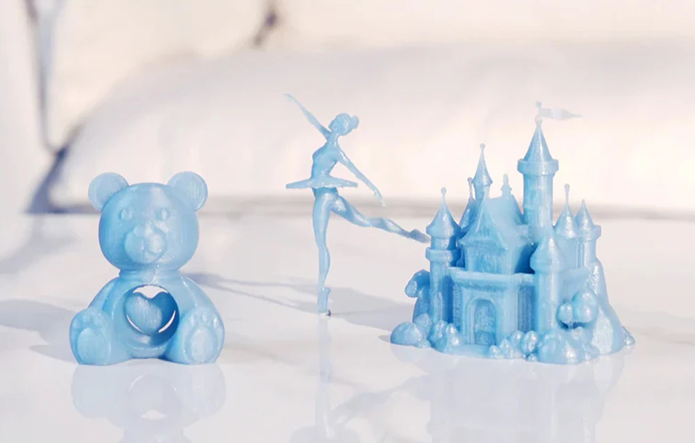 Imprimări 3D albastru lucios ale unui ursuleț de pluș, unei balerine și unui castel realizate din filament Twinkling PLA