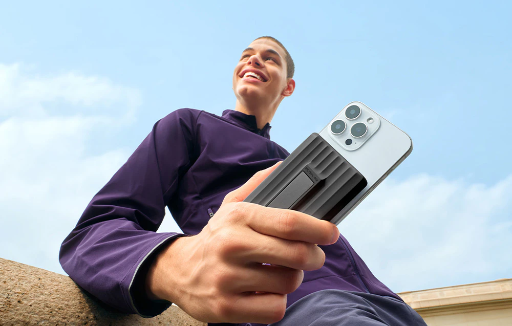 Un bărbat zâmbitor într-o jachetă mov stă sprijinit de un perete și ține un smartphone conectat la un powerbank magnetic.