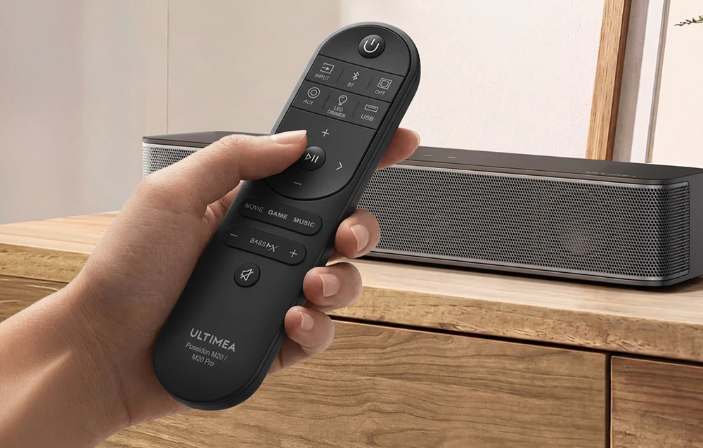 O telecomandă pentru soundbar, ținută în mână, cu soundbar-ul așezat pe o comodă din lemn în fundal