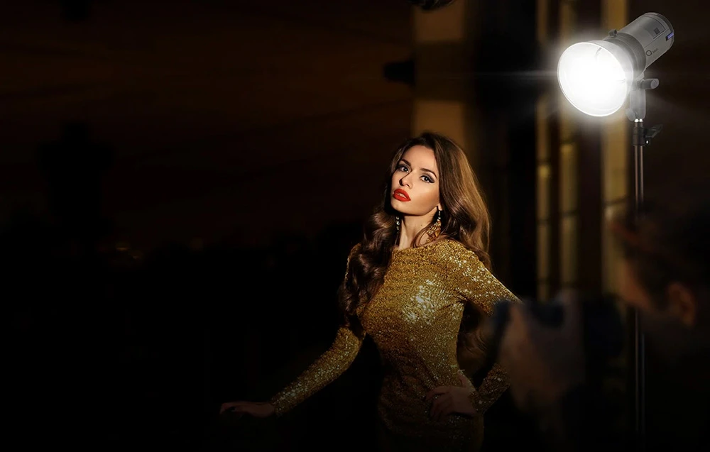 Un model într-o rochie aurie pozează noaptea, iluminată de lumina puternică a studioului din lateral.