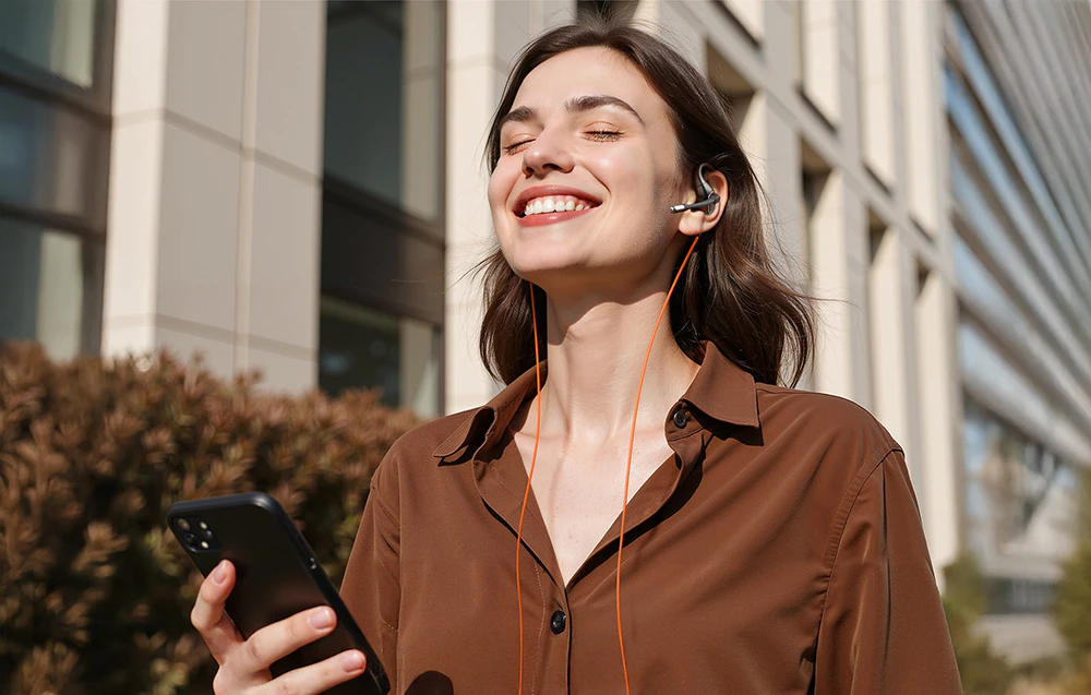 Femeie afară ținând telefonul în mână și zâmbind, purtând căști cu microfon