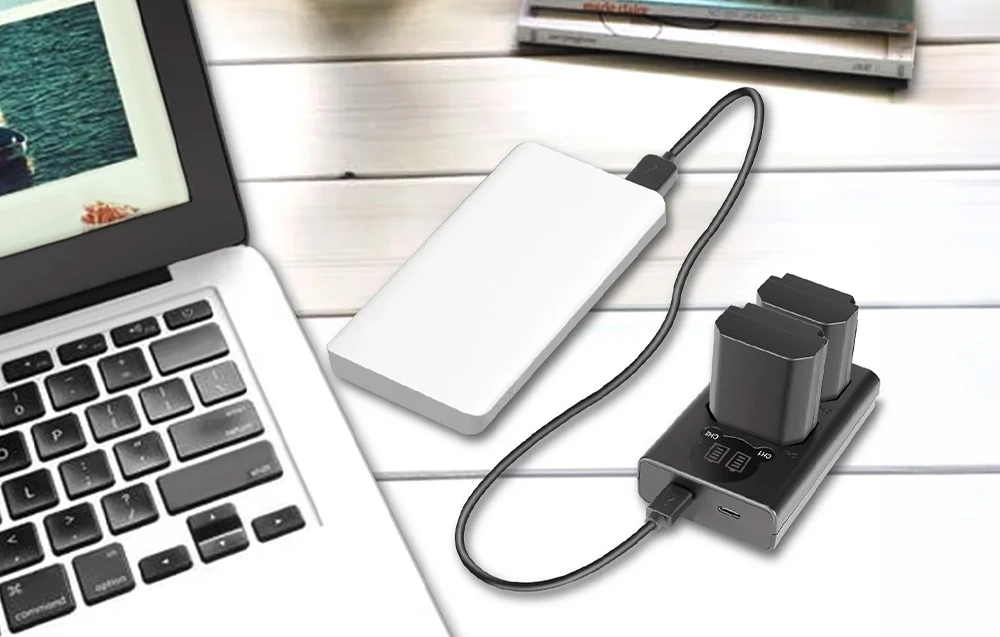 Un încărcător dublu pentru baterii conectat printr-un cablu USB la o baterie externă albă, lângă un laptop așezat pe o masă de culoare deschisă.