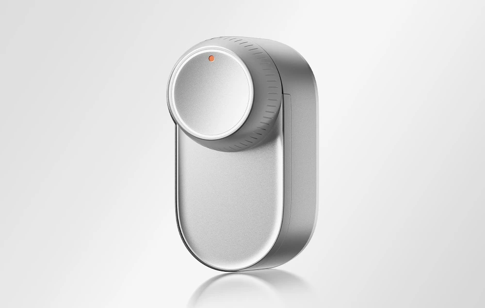 Încuietoare inteligentă SwitchBot în carcasă argintie cu buton rotund în partea de sus și indicator LED portocaliu