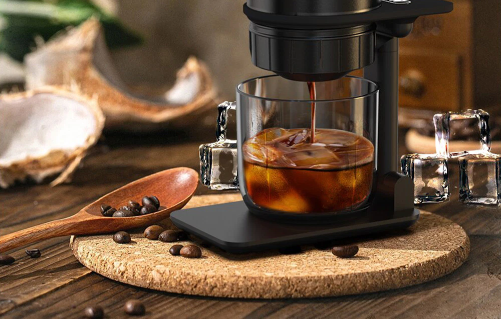 Cafeaua curge din aparatul de cafea într-un pahar umplut cu cuburi de gheață, așezat lângă boabe de cafea și cuburi de gheață.
