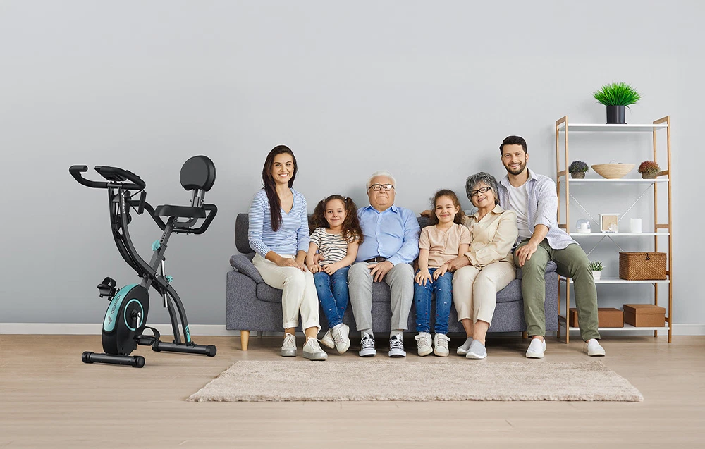 O familie multigenerațională așezată pe o canapea gri lângă o bicicletă de exerciții staționară MERACH, într-o cameră luminoasă