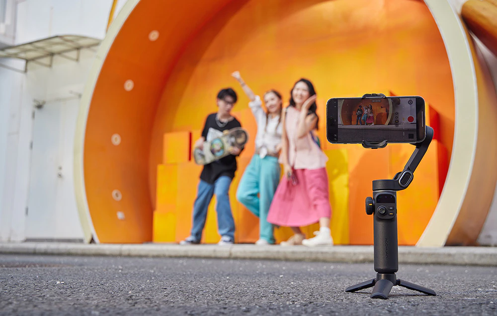 Un gimbal montat pe un mini-trepied, filmând trei prieteni care pozează fericiți pe o scenă portocalie.